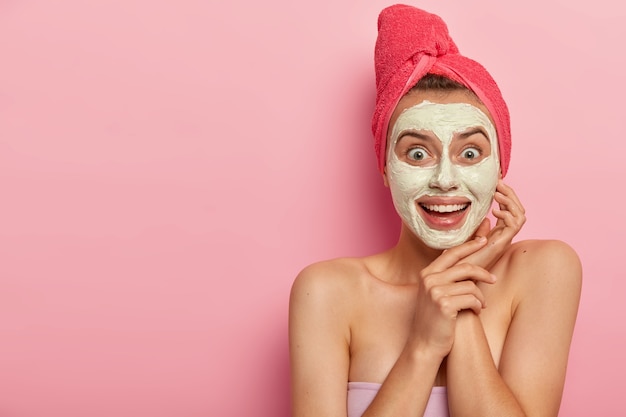 Happy joyful young woman applies cream mask wonderful for any skin type, wears red towel on head, nourishes skin, looks positively