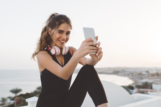 Happy joyful young woman in attractive sportswear making selfie on phone, smiling, enjoying sunrise in the morning on seafront. Cheerful mood, true happiness
