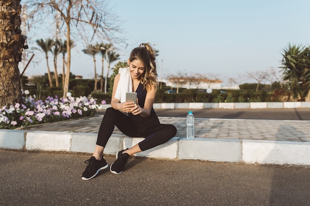 Happy joyful young woman in sportswear sitting outside on street of tropical city. Chatting on phone, expressing positivity, true emotions, healthy lifestyle, fitness, training