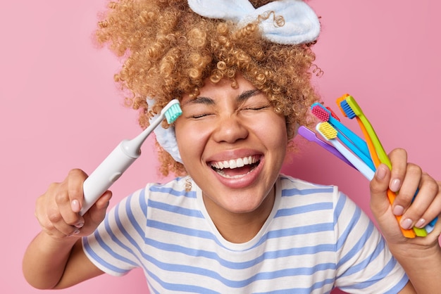 Felice gioiosa giovane donna con i capelli ricci tiene spazzolini da denti sorride ampiamente ha denti bianchi dopo la pulizia regolare indossa una maglietta a righe casual tiene gli occhi chiusi isolati su sfondo rosa