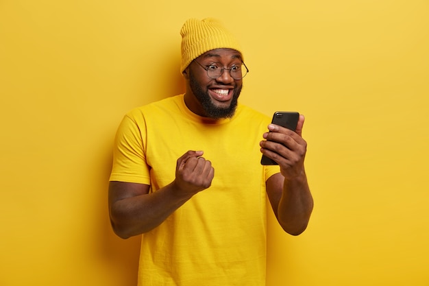 Happy joyous man clenches fist with triumph, watches football match online, focused in smartphone device, wears round spectacles