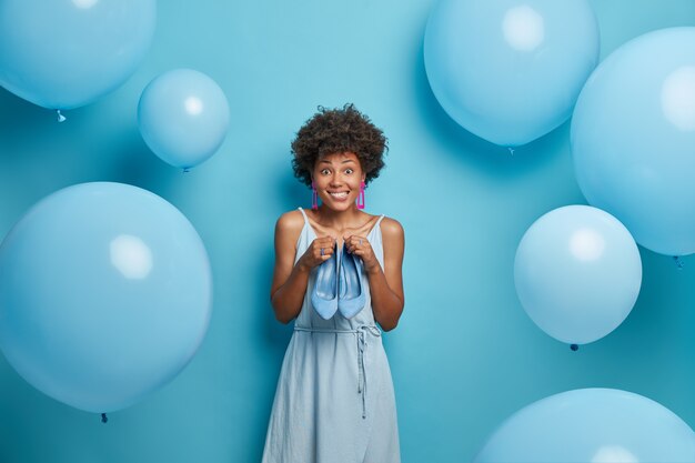 ピンクのドレスを着て ハイヒールの靴を持って 誕生日パーティーの準備をし 何を着るかを選択しようとし 脇を見て 青い風船の周りのアフロの髪のスタイリッシュな嬉しい女性の屋内ショット 無料の写真