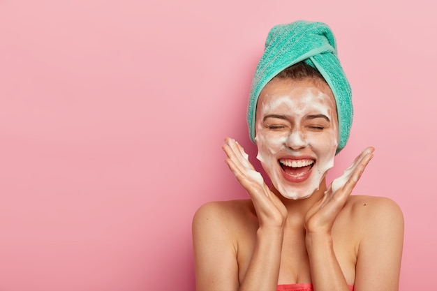 Happy joyous young girl spreads palms over face, washes face with soap, has fun in bathroom, pampers skin, wears wrapped towel on head, expresses positive emotions