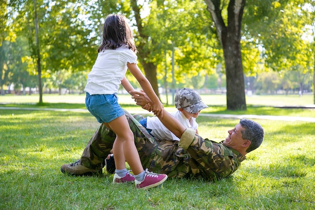 Happy kids and their military dad lying and playing on grass in park. Girl pulling fathers hand. Family reunion or returning home concept