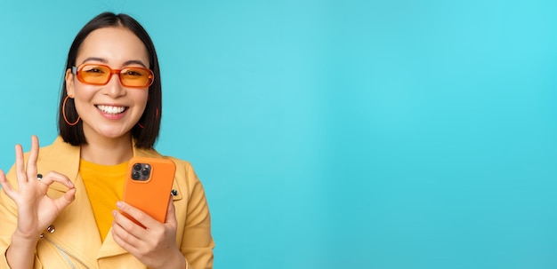 Happy korean girl in sunglasses showing okay sign and holding mobile phone using smartphone app recommending application standing over blue background