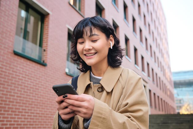 Happy korean girl walks on street listens music in wireless earphones and holds smartphone picks son