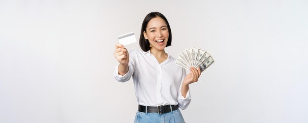 白いスタジオの背景にポーズをとって笑って笑ってクレジットカードとお金のドルを保持している幸せな韓国の女性