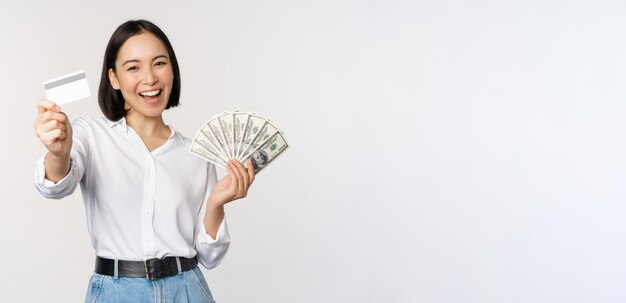 Happy korean woman holding credit card and money dollars smiling and laughing posing against white studio background