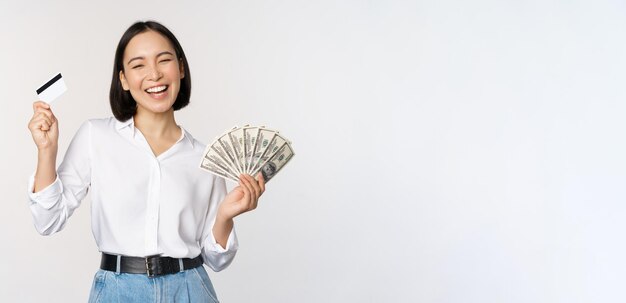 Счастливая кореянка с кредитной картой и деньгами в долларах улыбается и смеется, позируя на фоне белой студии