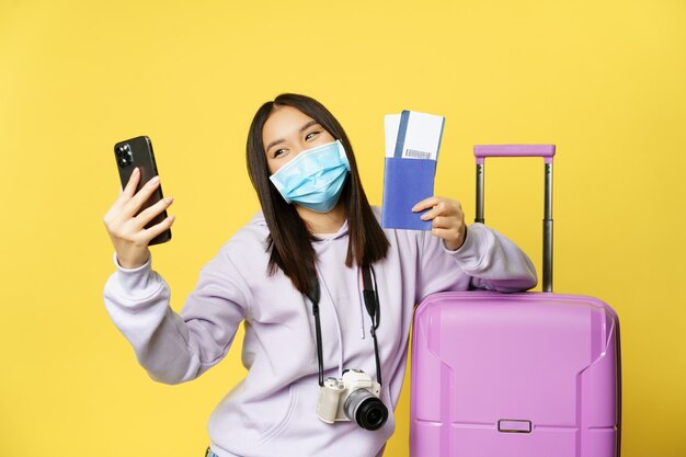 Счастливая корейская женщина, делающая селфи с паспортом и авиабилетами, собирается в отпуск, стоя возле би ...