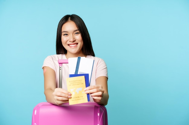 Happy korean woman, tourist with suitcare shows her health passport and tickets, covid-19 international vaccination certificate for travelling, going on trip, blue background.