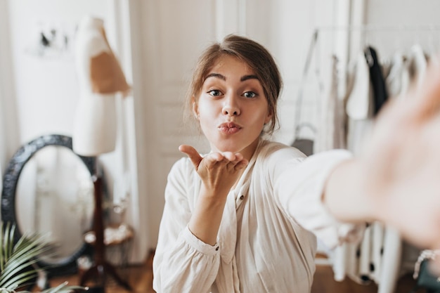 Happy lady in beige outfit blows kiss and takes selfie in office Attractive woman in white blouse poses in good mood in apartment