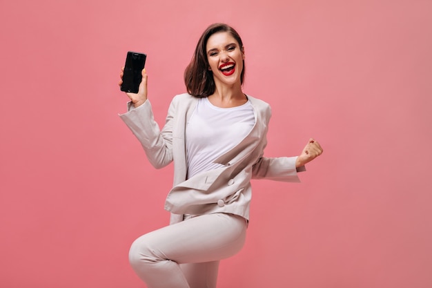 Счастливая дама в бежевом костюме позирует с телефоном на розовом фоне. Радостная девушка в офисном платье и с красными губами держит смартфон.