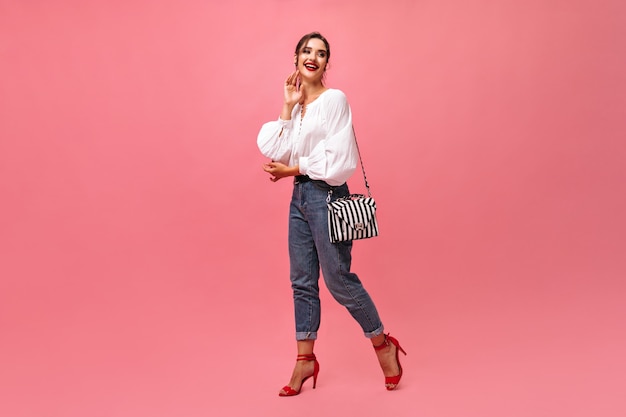 happy lady in jeans and white blouse posing on pink background.  smiling brunette with red lipstick and in modern outfit moves..
