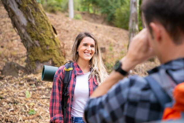 森の中の道路でポーズと笑顔の幸せな女性。彼のガールフレンドの写真を撮っている認識できない男。森の中で一緒にハイキングして楽しんでいる観光客。観光、冒険、夏休みのコンセプト