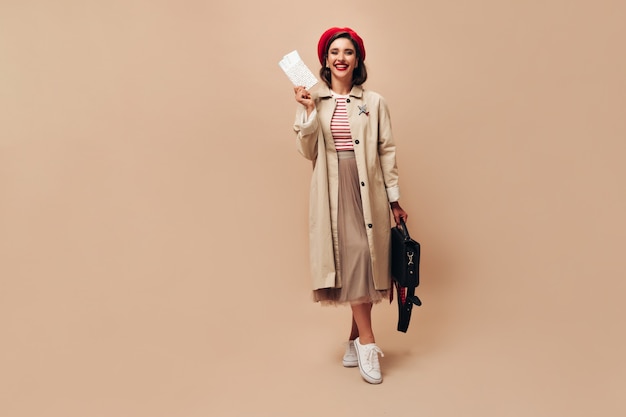 Happy Lady on red beret and trench poses with tickets on beige background. Joyful young woman in long stylish skirt and autumn coat looks at camera.