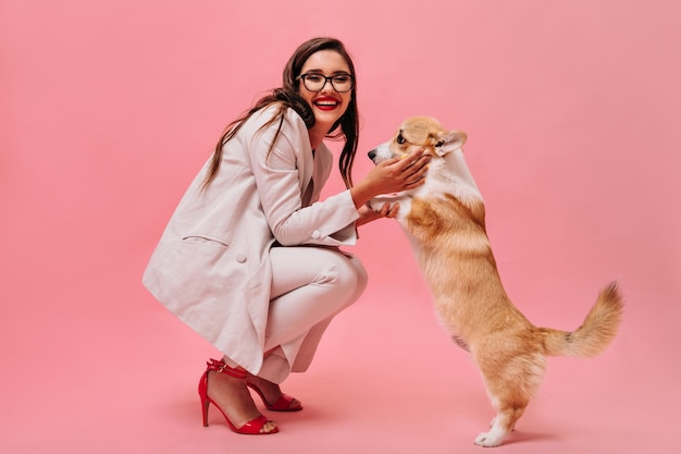 彼女の犬と一緒に楽しんでいるスーツと眼鏡の幸せな女性。明るくファッショナブルな服を着たうれしそうな女性は、孤立した背景にかわいいコーギーで遊ぶ。