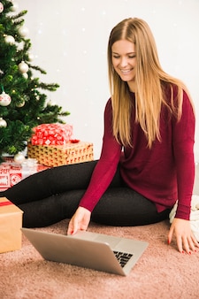 Happy lady using laptop near gift boxes and christmas tree