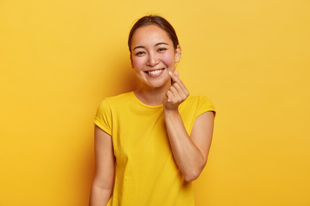Счастливая женщина с азиатской внешностью делает корейский знак, одетая в повседневную желтую футболку, с дружелюбным выражением лица стоит в помещении. Монохромный снимок. Язык тела. Женщина выражает любовь жестом