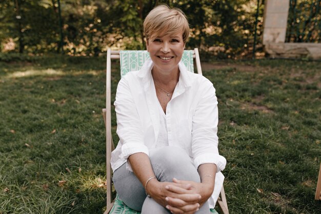 Happy lady with modern blonde hairstyle in light trendy blouse and trousers, smiling, looking into camera and sitting outdoor.