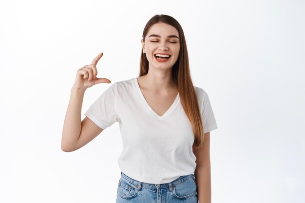 Happy laughing girl showing small size, little hand gesture and smiling, standing joyful against white background in casual clothes