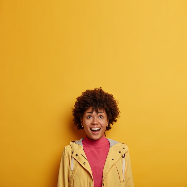행복한 웃는 여자는 관심과 기쁨으로 위를보고 캐주얼 재킷을 입은 재미있는 광고를 확인하고 곱슬 머리를 가지고 있으며 노란색 공간에 대해 포즈를 취합니다.