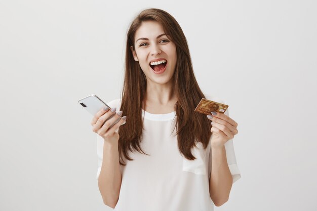 Happy laughing woman order online via smartphone app, holding credit card and mobile phone