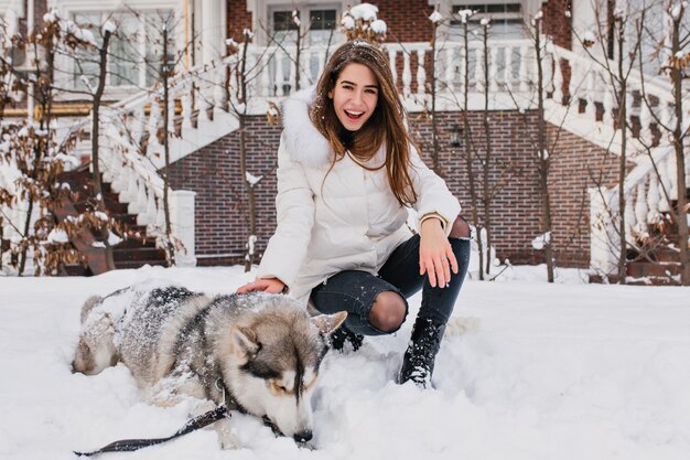 Happy laughing woman with straight hair sitting on snow beside her dog. Good-looking woman in jeans and white jacket posing with husky after walk in winter morning.