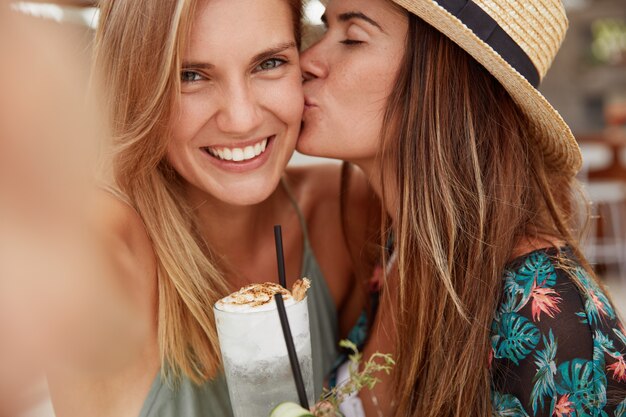 Happy lesbian females make selfie, kiss passionately, hold fresh cocktails, demonstrate good relationships and real love. Two female tourists enjoy summer journey, have summer party together