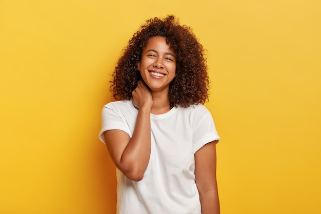 Happy lifestyle concept. Pleasant looking funny Afro woman feels lucky and satisfied, laughs happily, has white teeth with small gap, enjoys awesome day off, stands against yellow wall