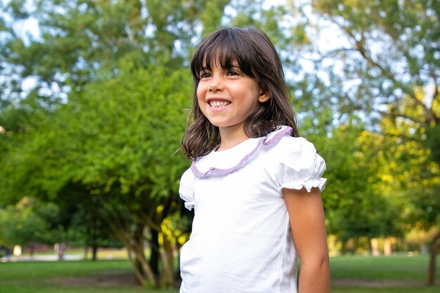 都市公園に立って、目をそらして笑っている幸せな小さな黒い髪の少女。夏の屋外で余暇を楽しむ子供。ミディアムショット。子供の頃の概念
