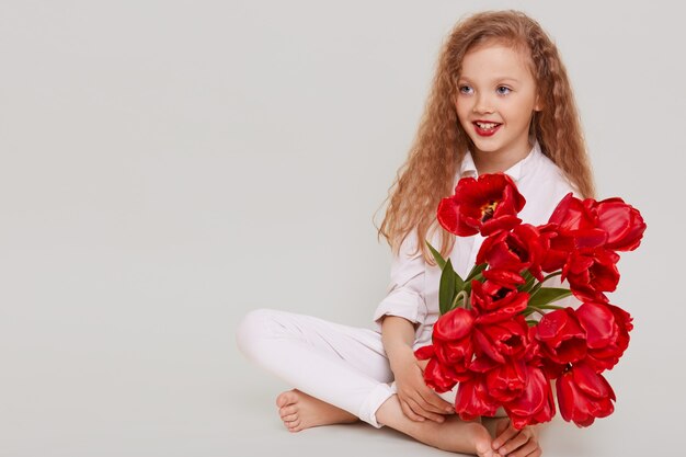 Happy little blonde girl looking away with toothy smile and positive facial expression, sitting on floor, holding bouquet of red tulips