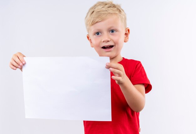 A happy little cute blonde boy in red t-shirt smiling and showing blank sheet of paper on a white wall