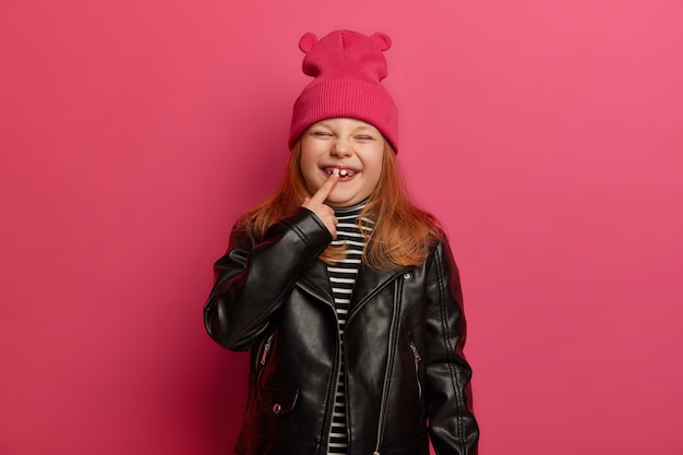 fashionabe 옷을 입은 행복 한 작은 생강 소녀는 그녀의 새로운 치아에서 잊을 수없는 어린 시절을 보내고 기쁨으로 얼굴을 가늘게 뜨고 분홍색 벽에 포즈를 취합니다. Chidren 성장 개념