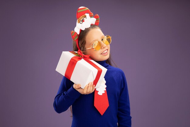 Happy little girl in blue turtleneck with red tie and  funny christmas rim on head holding a present looking intrigued standing over purple wall