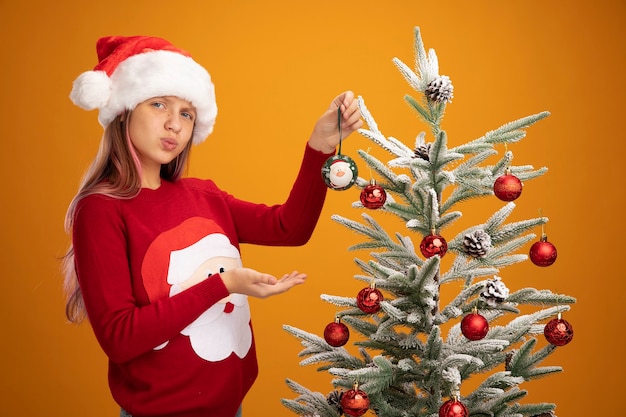 크리스마스 스웨터와 산타 모자에 행복 한 어린 소녀 팔을 제시 크리스마스 트리에 공을 매달려 오 오렌지 배경 위에 자신감을 찾고 그녀의 손