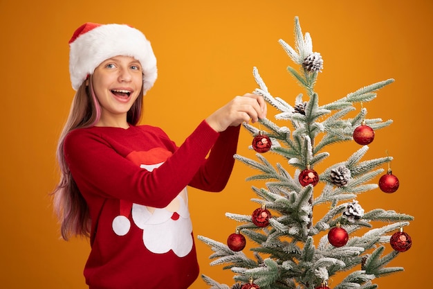 オレンジ色の背景の上で元気に笑ってクリスマスツリーにボールをぶら下げてクリスマスセーターとサンタ帽子の幸せな少女
