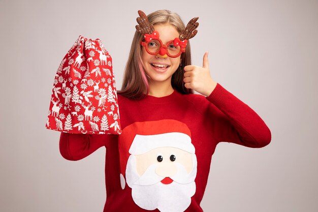흰색 배경 위에 서 엄지 손가락을 유쾌 하 게 보여주는 미소를 카메라를보고 선물 산타 빨간 가방을 들고 재미있는 파티 안경을 쓰고 크리스마스 스웨터에 행복 한 어린 소녀