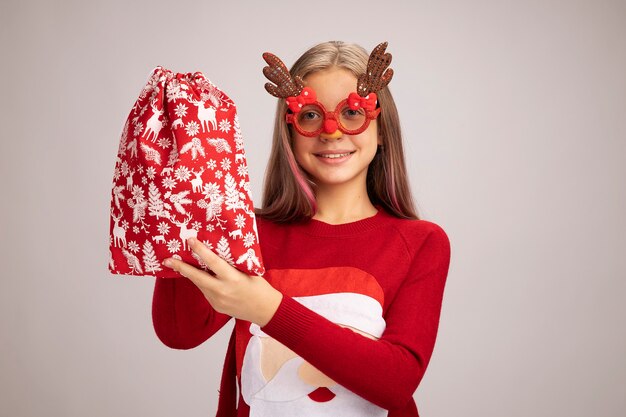 Happy little girl in christmas sweater wearing funny party glasses holding santa red bag with gifts looking at camera smiling cheerfully standing over white background