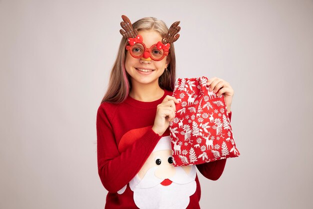 Счастливая маленькая девочка в рождественском свитере в забавных партийных очках, держащая санта-красный мешок с подарками, глядя в камеру, весело улыбаясь, стоя на белом фоне