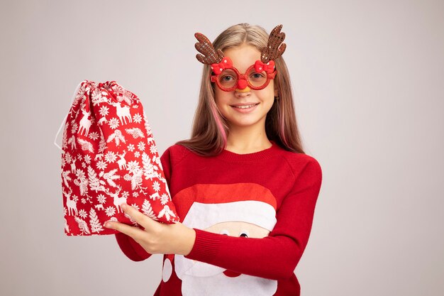 Happy little girl in christmas sweater wearing funny party glasses holding santa red bag with gifts looking at camera smiling cheerfully standing over white background