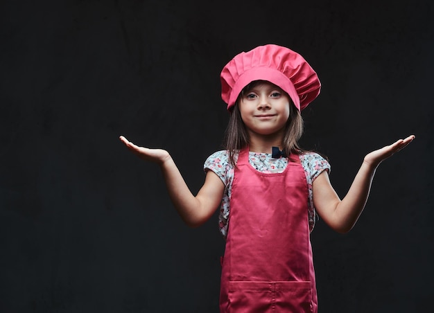 Free photo happy little girl dressed in pink cook posing in a studio. isolated on dark textured background.