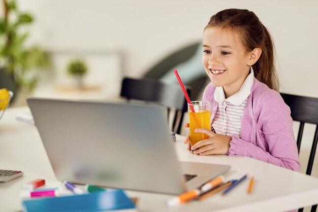 집에서 노트북을 통해 학습하는 동안 주스를 마시는 행복한 어린 소녀