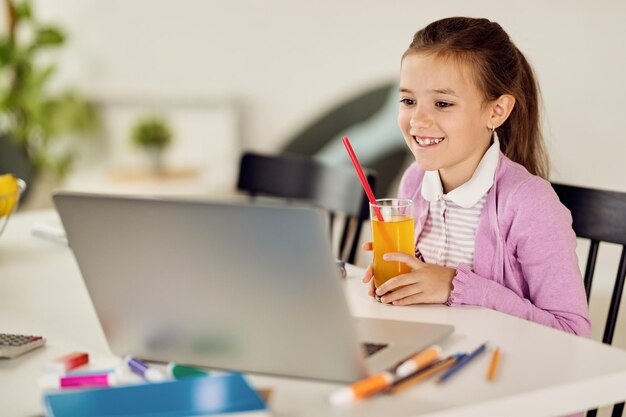 Happy little girl drinking juice while elearning over laptop at home