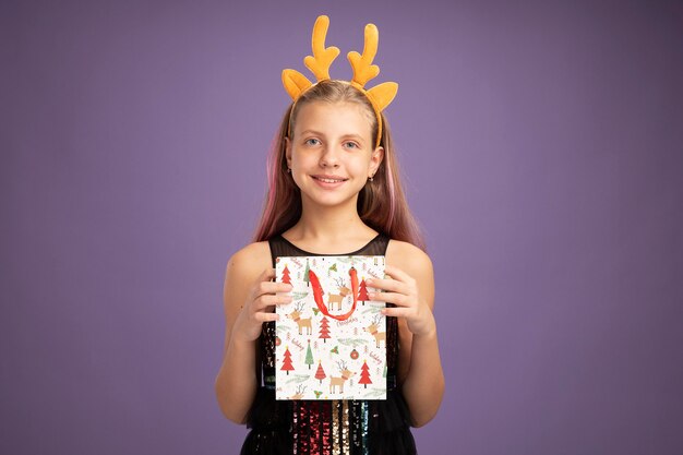 Happy little girl in glitter party dress and funny headband with deer horns holding christmas paper bag with gifts looking at camera smiling cheerfully standing over purple background