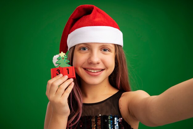 Happy little girl in glitter party dress and santa hat holding toy cubes with new year date looking at camera smiling cheerfully standing over green background