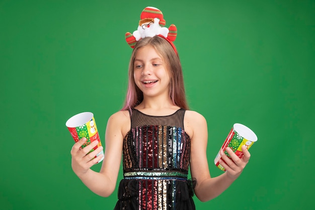 Free photo happy little girl in glitter party dress and santa headband holding two colorful paper cups smiling cheerfully standing over green background