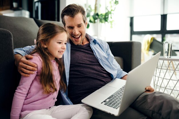 Happy little girl and her father using a computer and watching something on the internet while relaxing in the living room