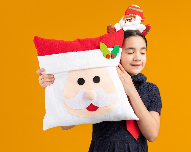 닫힌 된 눈으로 크리스마스 베개를 들고 머리에 재미있는 테두리와 빨간 넥타이를 착용하는 니트 드레스에 행복 한 어린 소녀 미소