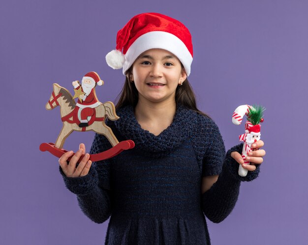 Happy little girl in knit dress wearing santa hat holding christmas toys  smiling cheerfully  standing over purple wall